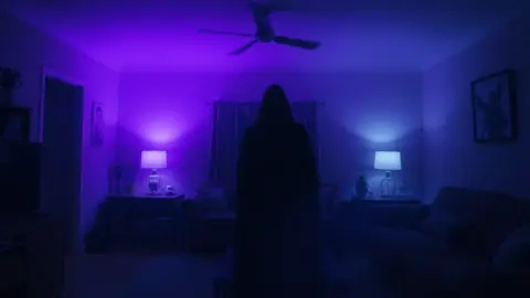 Dim living room with several faint overlapping silhouettes of a witch in different spots