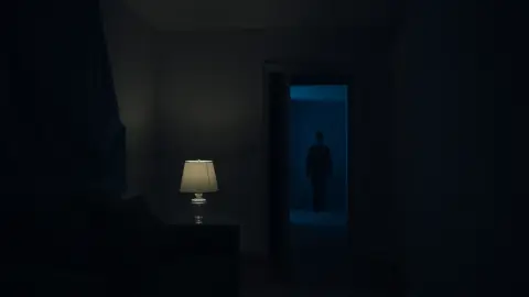Dim living room at night with a shadowy figure standing in the hallway doorway