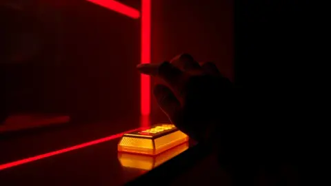 Hand reaching toward a glowing gold bar between thin red laser beams