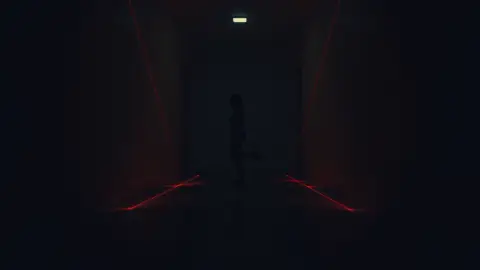 Person tiptoeing on one leg through a dim hallway between thin red laser beams