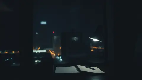 Desk lamp glowing in a dark apartment at night with city lights outside